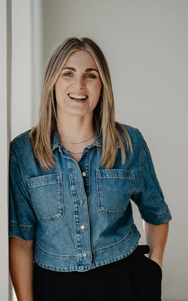 Sarah Segerer lehnt mit verschränkten Armen an der Wand. Sie lächelt freundlich und trägt ein schwarzes T-Shirt.