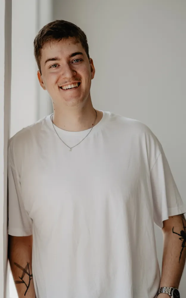 Simon Henschel lehnt an der Wand. Sie lächelt freundlich und trägt ein weißes T-Shirt.